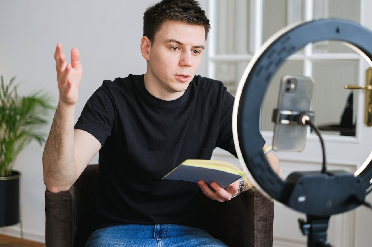 Young man in black t-shirt vlogging indoors using a smartphone and ring light.